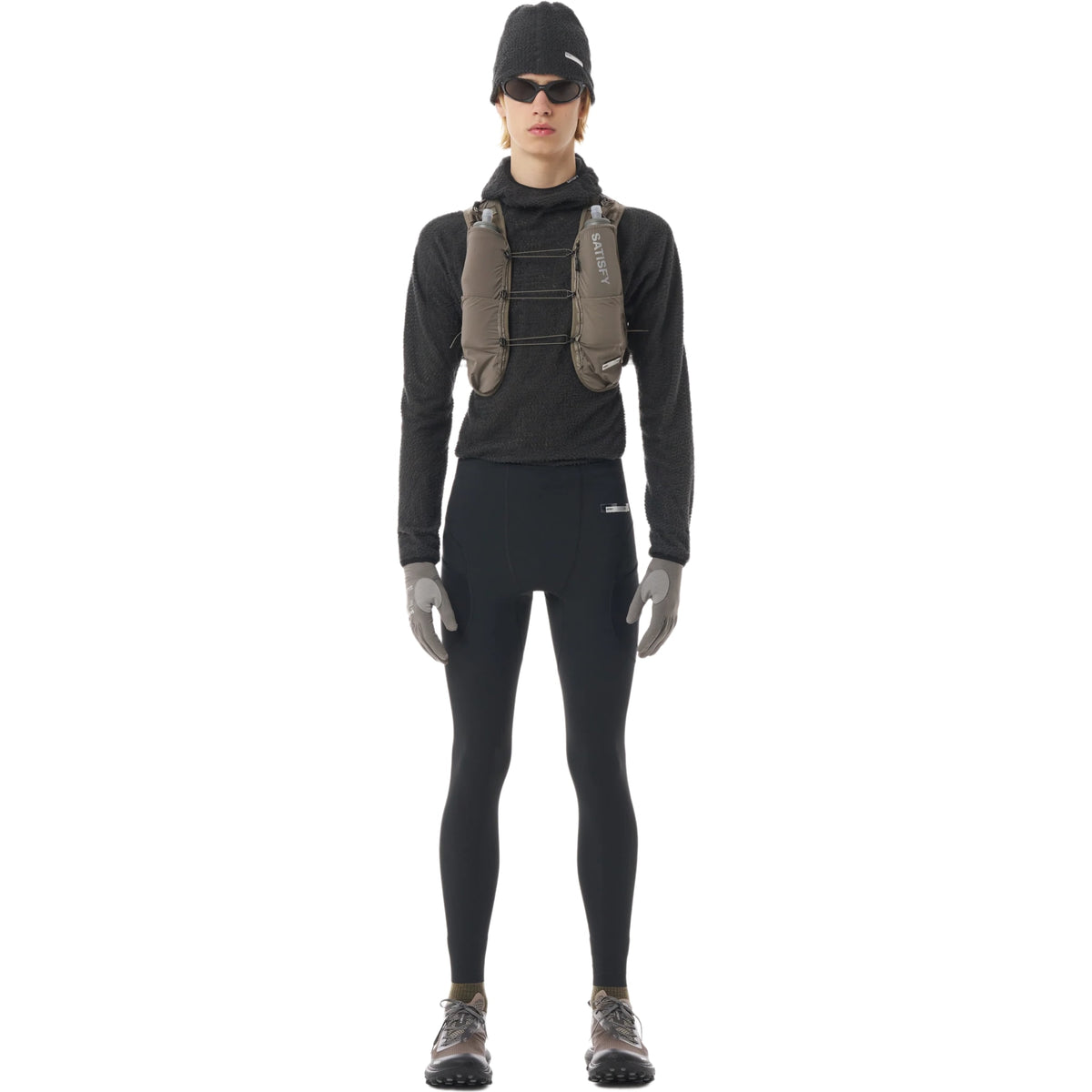 A person facing forward against a plain white background wearing black Satisfy TechSilk Evo Cargo Tights, along with other athletic gear including a hydration vest, cap, sunglasses, gloves, and running shoes.