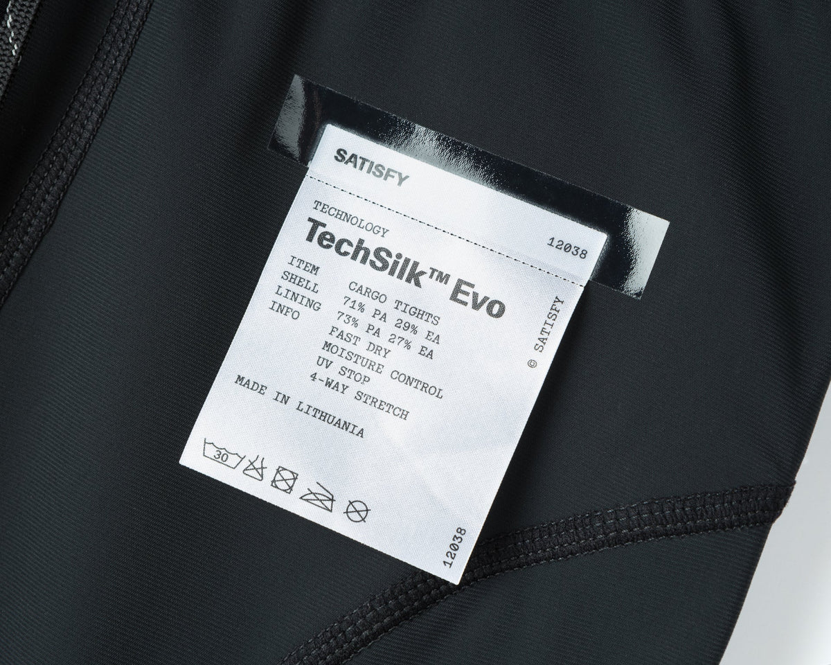 A close-up of a Satisfy clothing label reading TechSilk Evo, detailing materials, wash symbols, and stating the item is made in Lithuania. The label is sewn onto black fabric.