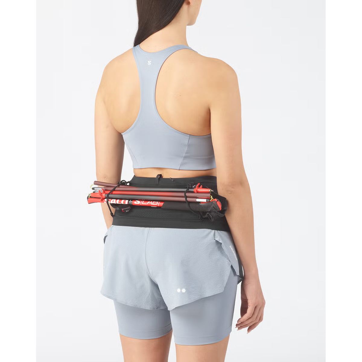 A woman in a light blue sports outfit faces away, wearing a Salomon ADV Skin Seamless Belt Black that holds folded trekking poles.