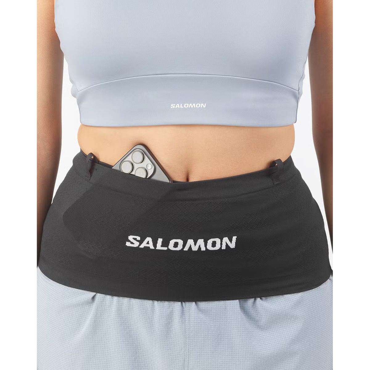 A person shown from the waist down, dressed in a light blue sports top and shorts, wears the Salomon ADV Skin Seamless Belt Black running belt with a phone partially tucked inside.