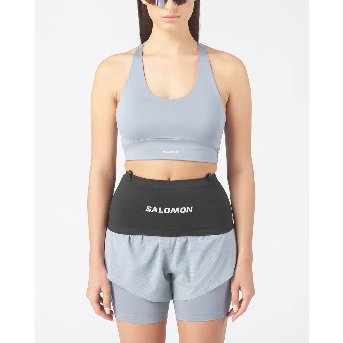 A woman stands facing forward against a plain white background, wearing a light gray sports bra, matching shorts, and the Salomon ADV Skin Seamless Belt in black.