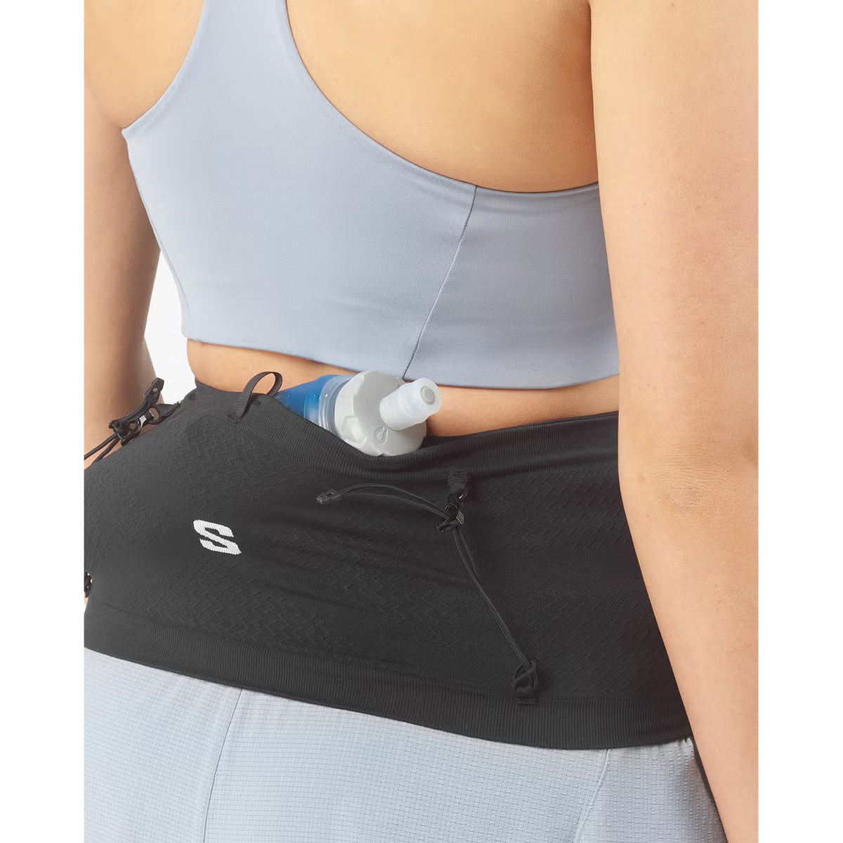 A person dressed in athletic wear is seen from behind with a Salomon ADV Skin Seamless Belt Black, carrying a soft water bottle inside. They are wearing a light gray sports bra and leggings.
