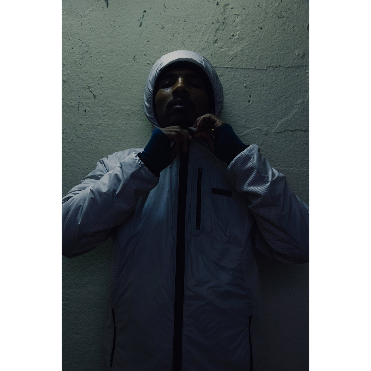 A person in a white hooded jacket zips up the front, standing against a textured wall in low, moody lighting.