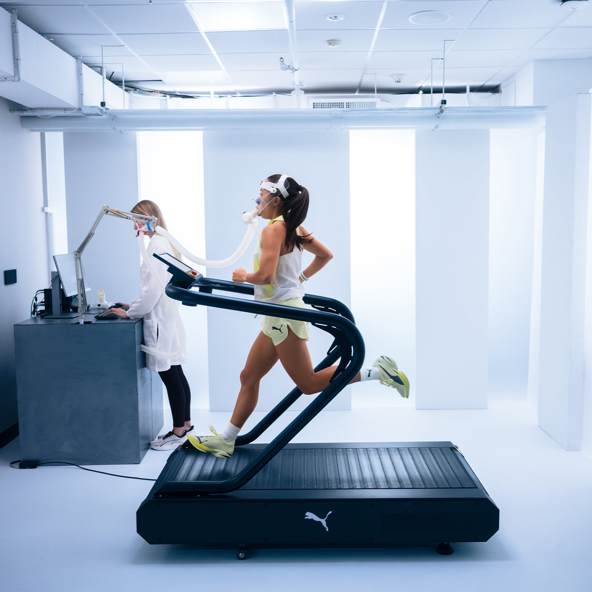 A woman runs on a treadmill wearing a mask connected to a machine, while a technician monitors data on a computer in a bright, modern lab.