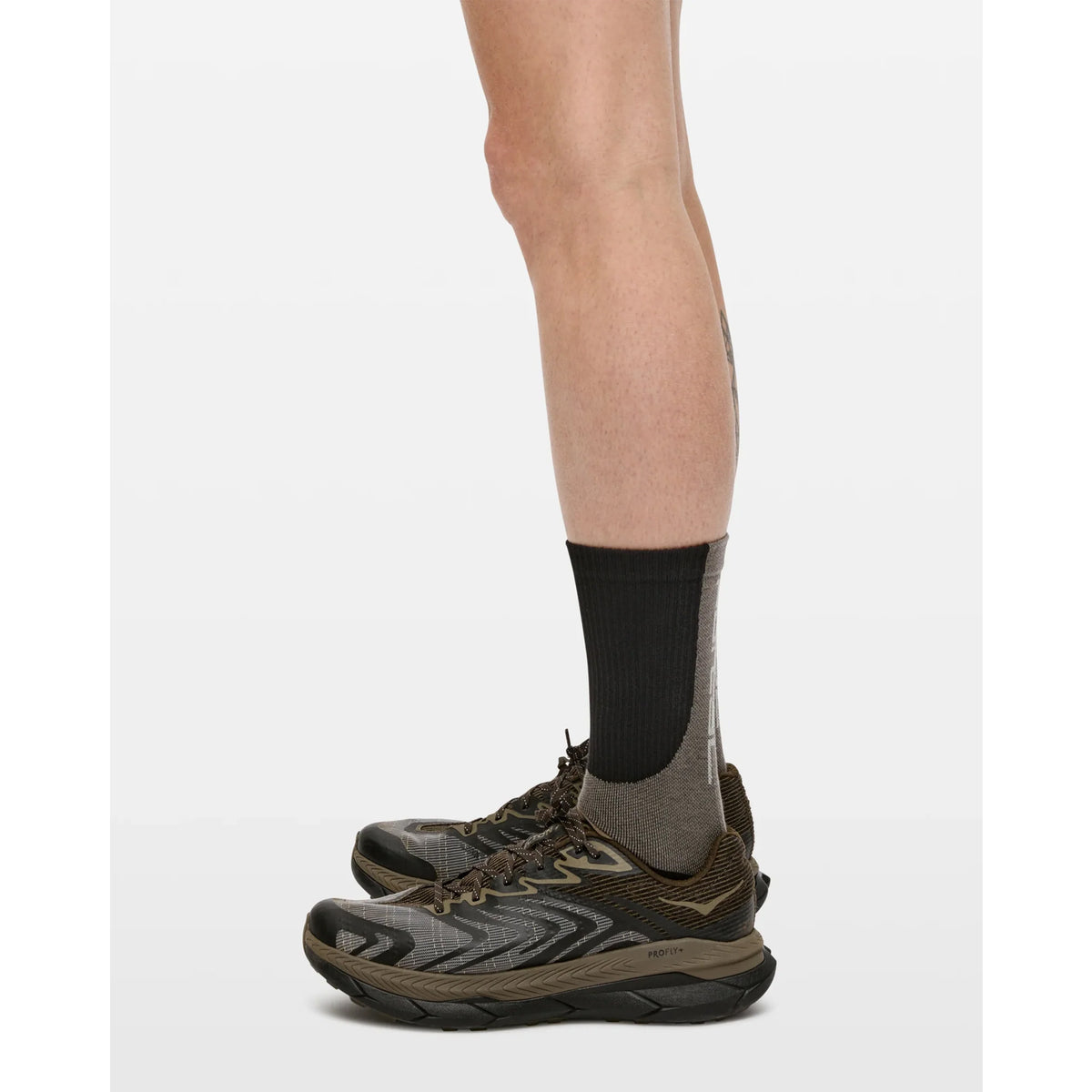 Side view of a person’s lower legs wearing Portal Lineage All Purpose Socks in Black Beauty with black and brown trail running shoes, standing on a plain white background.