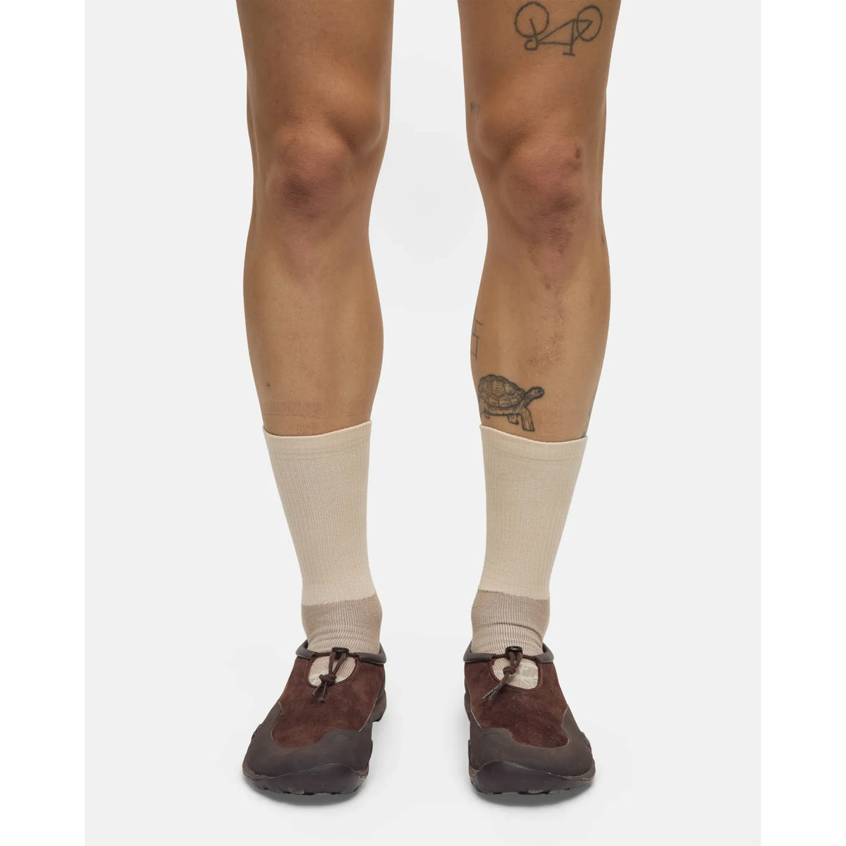 A person wearing Portal Lineage All Purpose Socks in Greige and brown shoes stands with their legs visible; they have tattoos on both legs, including a turtle and a bicycle.