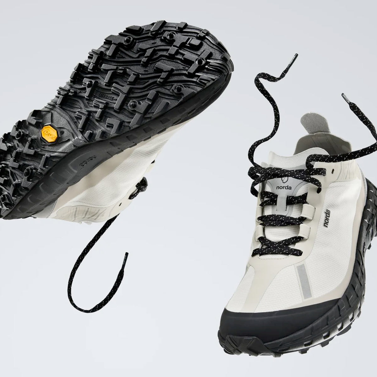 Two white trail running shoes with black soles and laces appear to float in midair. One shoe shows its rugged sole, while the other displays the top view. The background is plain and light-colored.