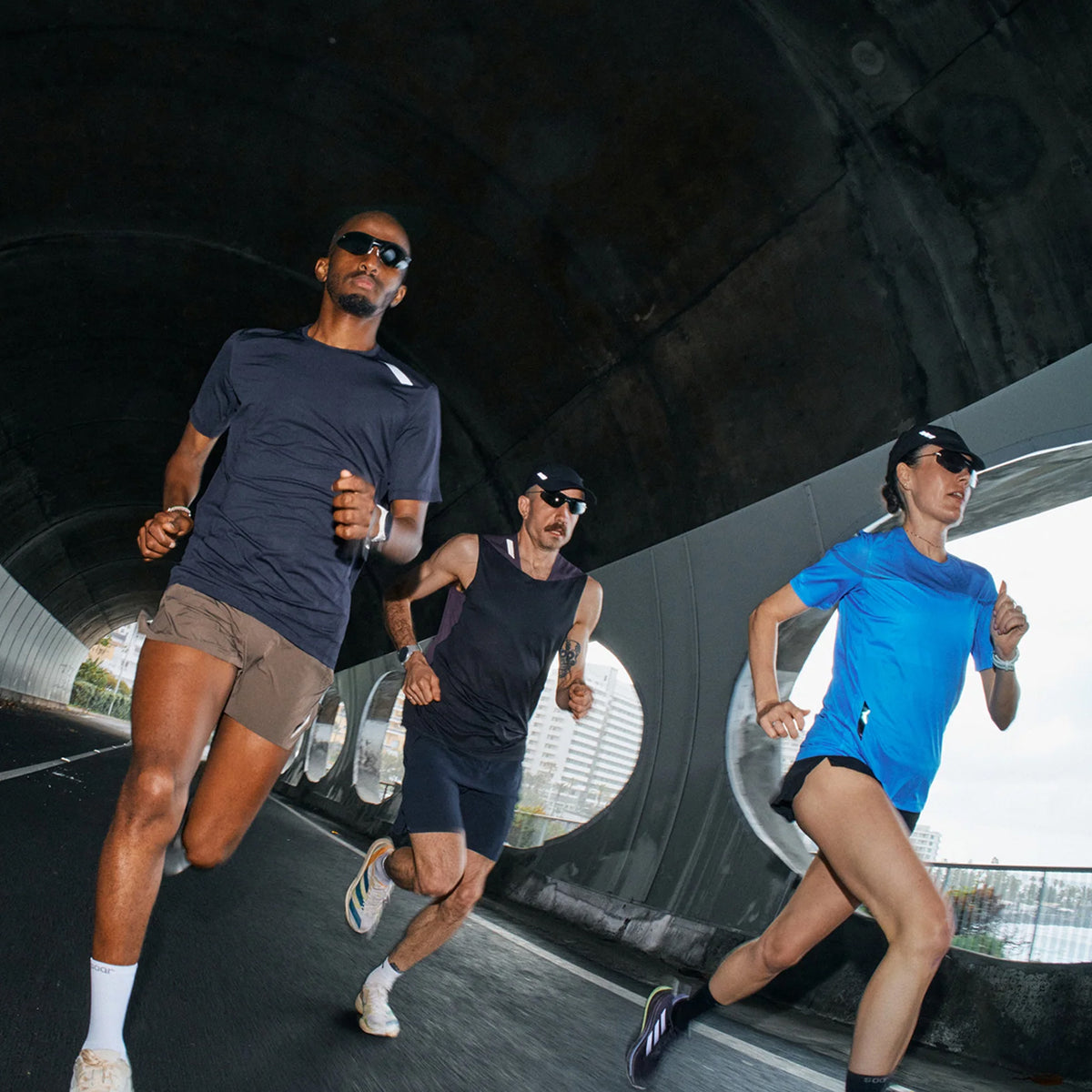 Three people in athletic gear run together inside a tunnel. Two men and one woman are wearing sunglasses and hats, running on a paved road with city buildings visible through round windows.