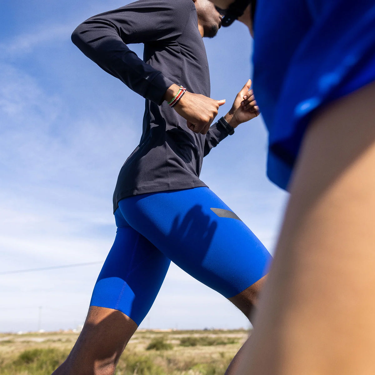 Two people running outdoors; the focus is on one runner wearing a black long-sleeve shirt and blue shorts. Another runner wearing blue is partially visible in the foreground. The background shows a clear sky and grassy landscape.