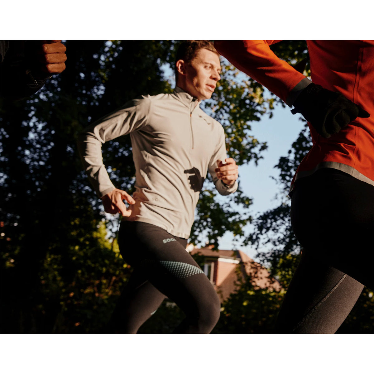 Several people are jogging outdoors in athletic wear, with sunlight filtering through trees and houses visible in the background.