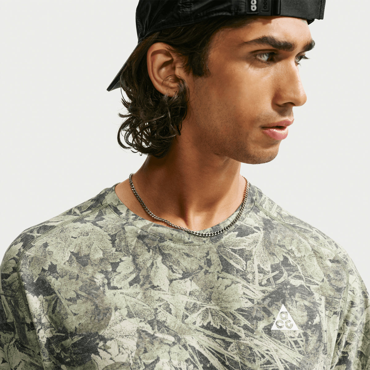 A person with long brown hair wears a backward black cap, a chain necklace, and a green leafy-patterned shirt with a triangle logo, looking to the side against a plain background.