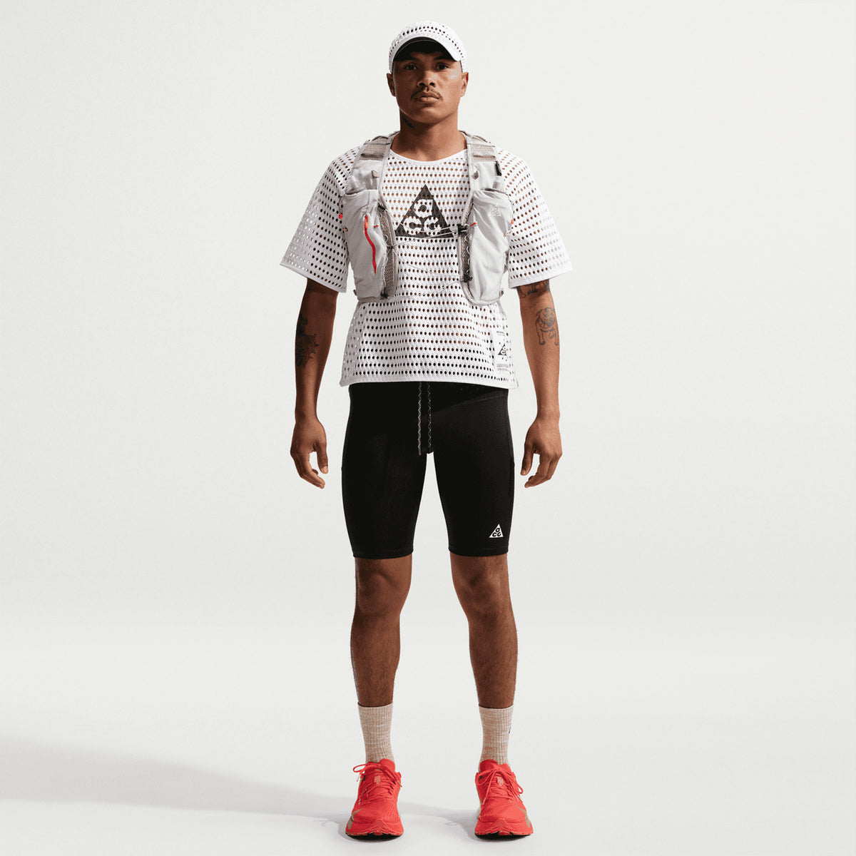 A person stands facing forward in a white cap, Nike Men’s ACG Radical AirFlow Tee White / Anthracite, gray vest, black cycling shorts, beige socks, and bright red sneakers against a plain light background.