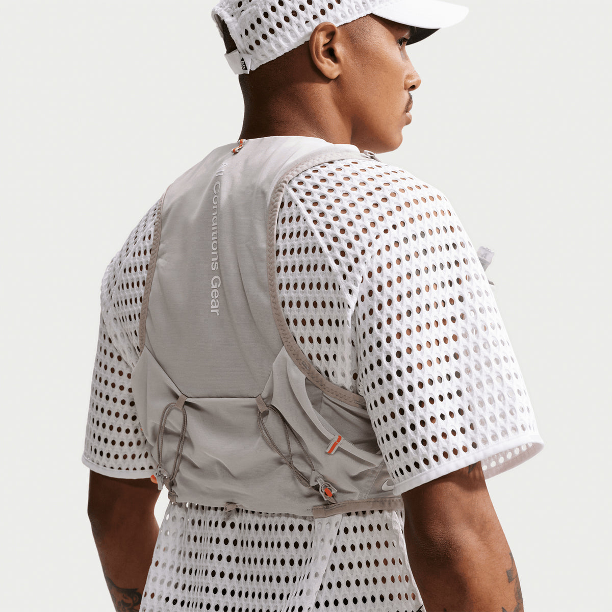 A person wearing the Nike Men’s ACG Radical AirFlow Tee White / Anthracite, a white cap, and a light gray vest with Conditions Gear text on the back, viewed from behind against a plain background.