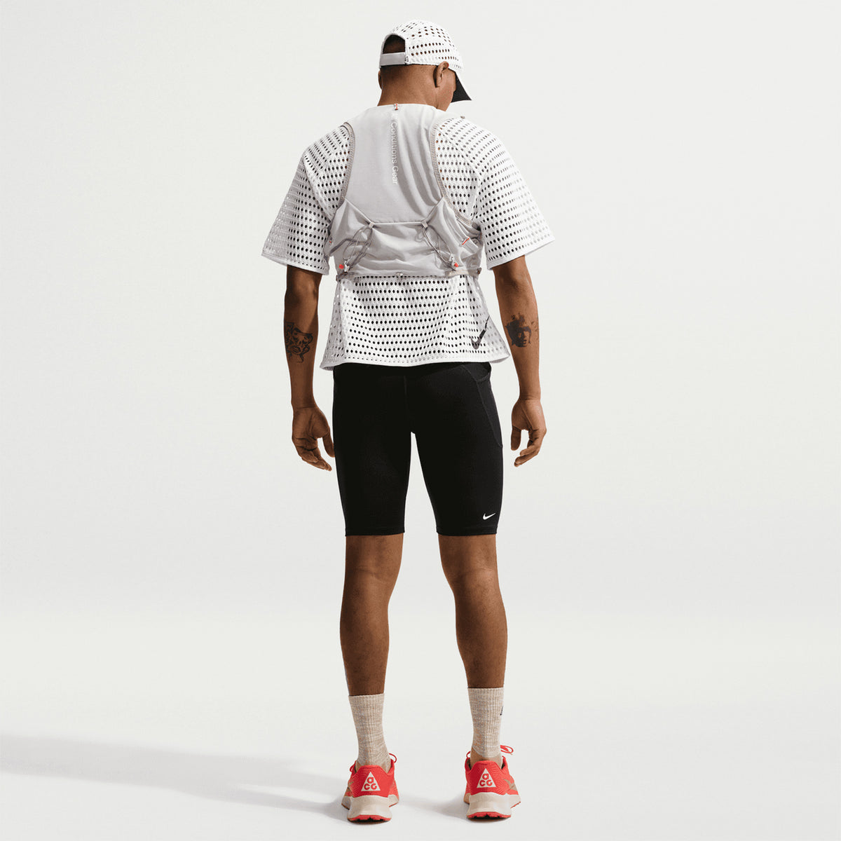 A person in a Men’s ACG Radical AirFlow Tee White / Anthracite by Nike, black shorts, light gray backpack, beige socks, bright red sneakers, and white cap stands facing away against a plain white background.