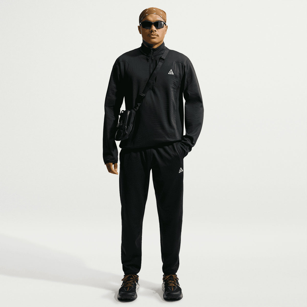 Clad in the Nike Men’s ACG Wolf Grid 1/2 Zip Top in Black / Summit White, along with a crossbody bag, sunglasses, and sneakers, a person stands facing forward against a plain white background—exuding both style and breathable warmth.
