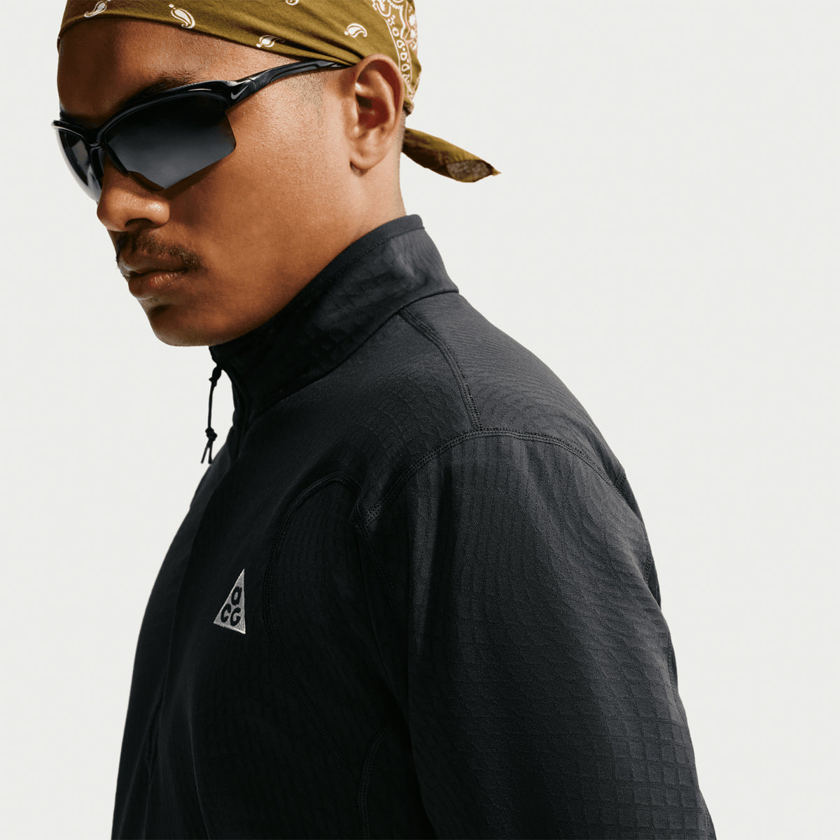A person wearing dark sunglasses, a brown bandana, and a black Nike Men’s ACG Wolf Grid 1/2 Zip Top in Black / Summit White with an ACG logo poses against a plain white background. The grid fleece top from Nike provides breathable warmth while maintaining a sleek and modern look.