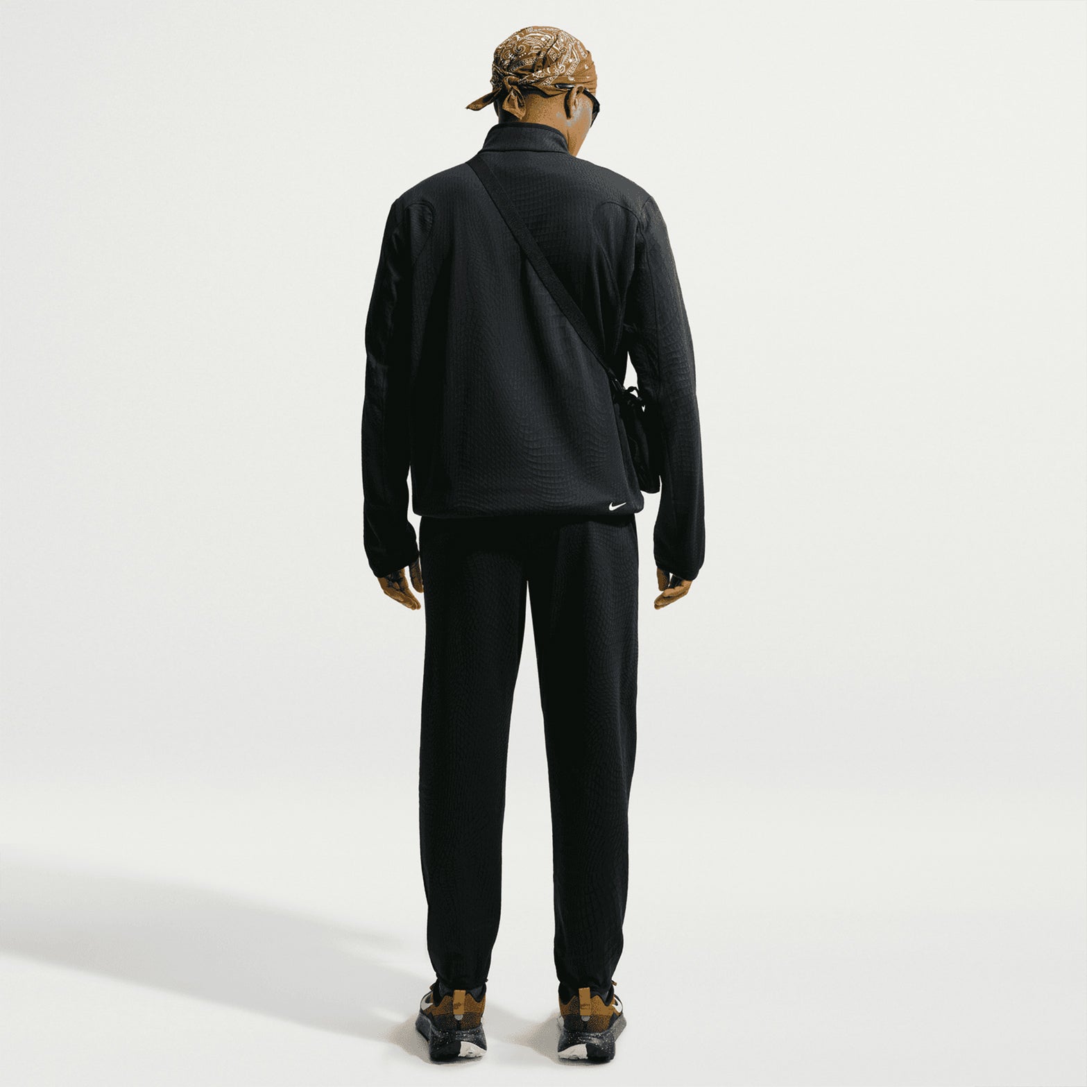 A person stands facing away, dressed in an all-black textured tracksuit featuring the Nike Men’s ACG Wolf Grid 1/2 Zip Top in Black / Summit White for breathable warmth. Their look is completed with black shoes, a brown patterned headscarf, sunglasses, and a crossbody bag. The background is plain and light-colored.