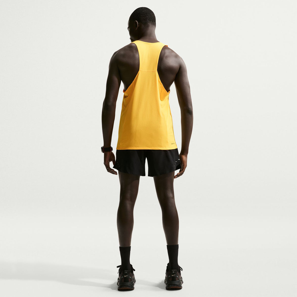 A person stands facing away, wearing a yellow sleeveless athletic top and Nike Men’s Dri-FIT Stride 2 In 1 7 Inch Shorts in black, paired with black socks and black shoes, against a plain white background.