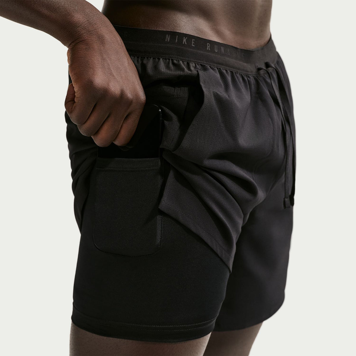 A person wearing Nike Men’s Dri-FIT Stride 2 In 1 7 Inch Shorts in black places a hand in a side pocket, set against a plain, light-colored background.
