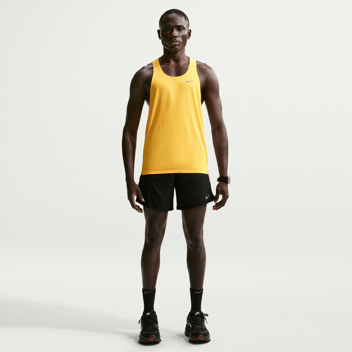 A person wearing a yellow tank top, Nike Men’s Dri-FIT Stride 2 In 1 7 Inch Shorts in black, black socks, and black sneakers stands facing forward against a plain white background.