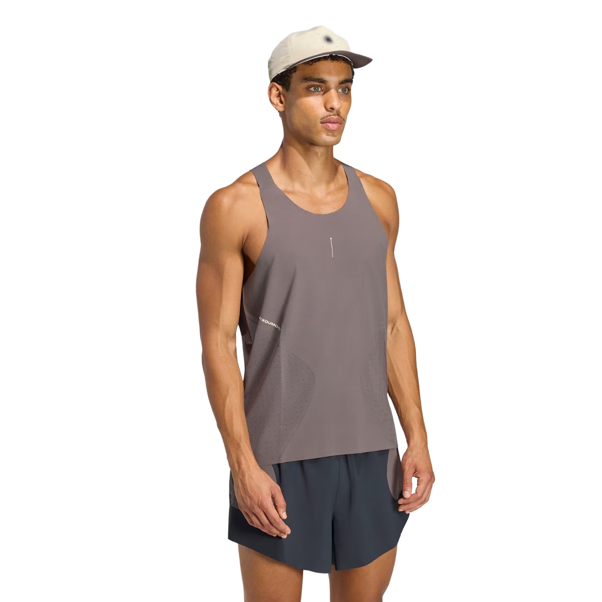 A man wearing an Adidas Men’s Hermanos Koumori Running Tank in charcoal, black shorts, and a light beige cap stands against a white background, ready for performance running.