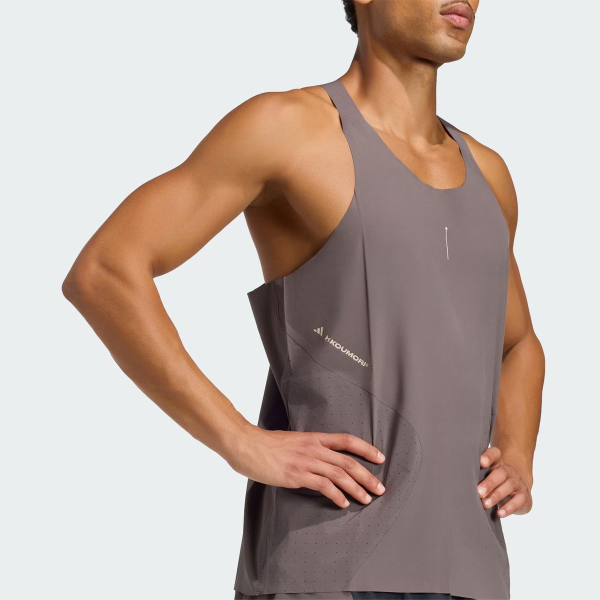 A person wearing a charcoal Men’s Hermanos Koumori Running Tank by Adidas stands with their hands on their hips, facing slightly to the side. The performance top features small perforations and a subtle logo near the side seam.
