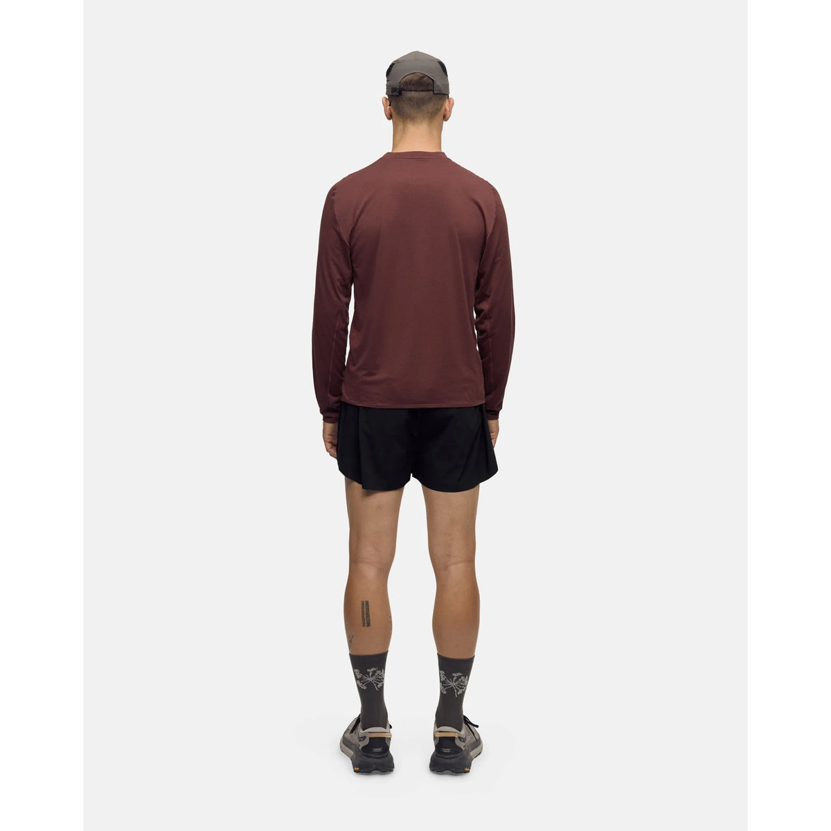 A person seen from behind wearing a Portal Men’s Lateral Peak Long Sleeve Top Bitter Chocolate, black shorts, patterned crew socks, and sneakers.