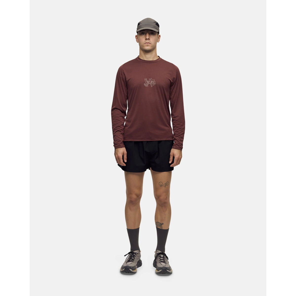 A person wears a dark brown Portal Men’s Lateral Peak Long Sleeve Top with a small graphic on the chest, black athletic shorts, gray socks, and sneakers.