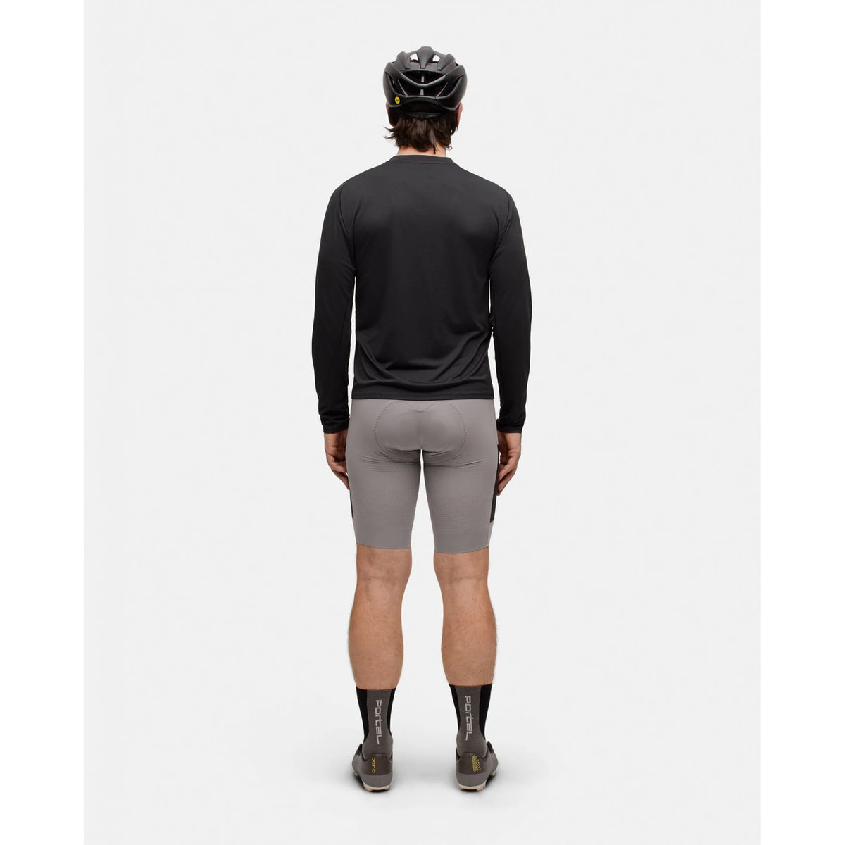A man wearing a black cycling helmet, Portal Men’s Lateral Peak Long Sleeve Top Black Beauty, gray cycling shorts, black socks, and cycling shoes stands facing away from the camera against a plain white background.