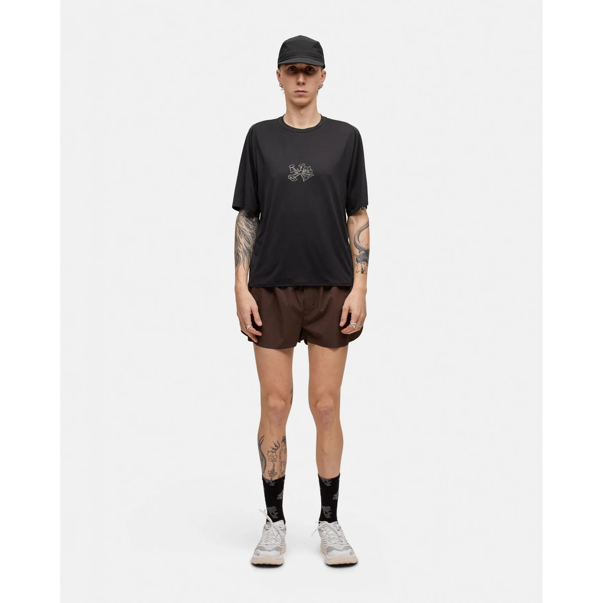 A person stands facing forward against a plain white background, wearing the Portal Men’s Lateral Peak Tee in Black Beauty—a loose, moisture-wicking black T-shirt with a small graphic—paired with a black cap, brown shorts, black socks, and light sneakers. Tattoos are visible on their arms and legs.