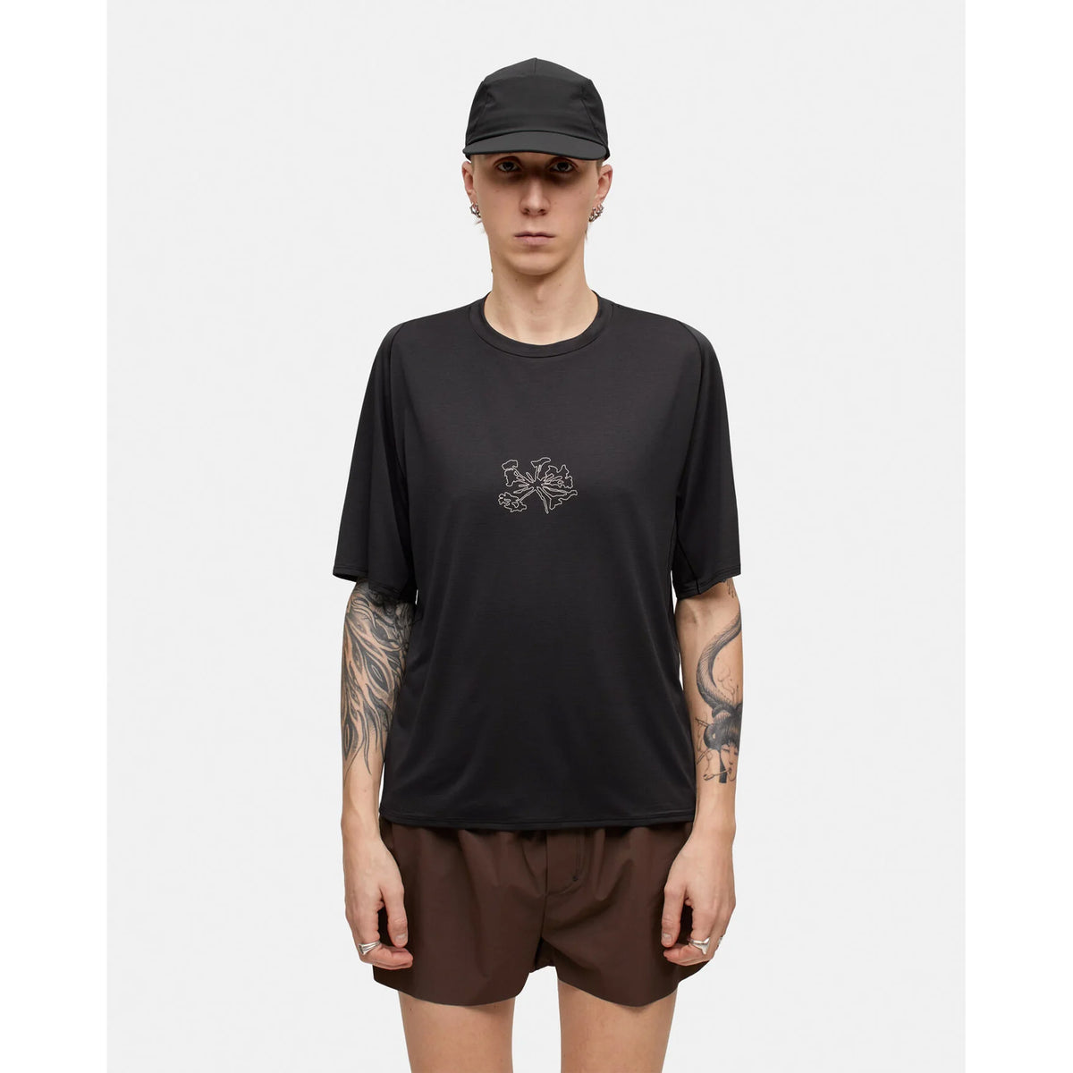 A person stands facing forward against a plain white background, wearing a black Portal Men’s Lateral Peak Tee Black Beauty with a small chest graphic, paired with a black cap and brown shorts.