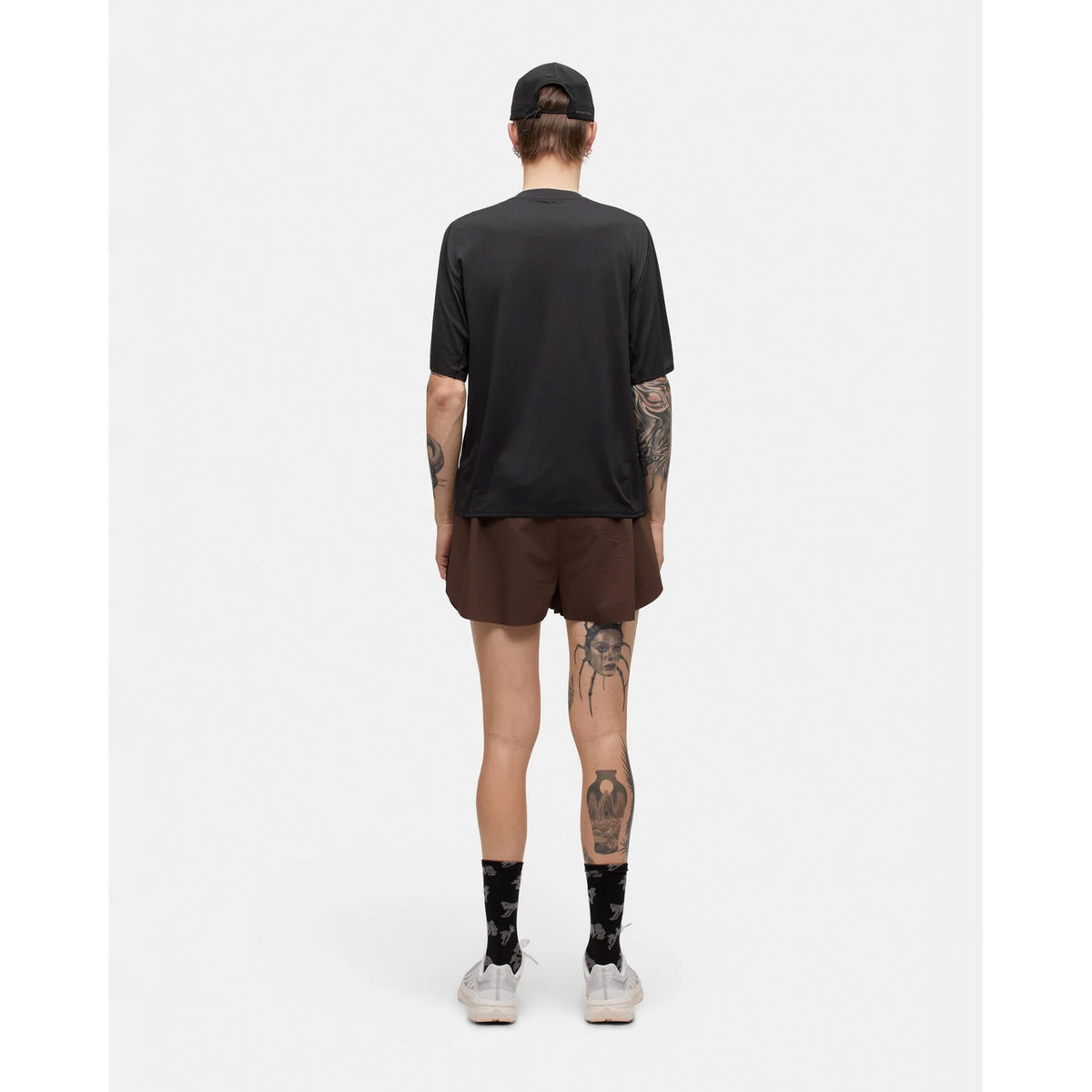 A person stands facing away against a plain white background, wearing the Portal Men’s Lateral Peak Tee in Black Beauty, paired with brown shorts, a black cap, patterned black socks, and white sneakers. Tattoos are visible on their arms and legs.