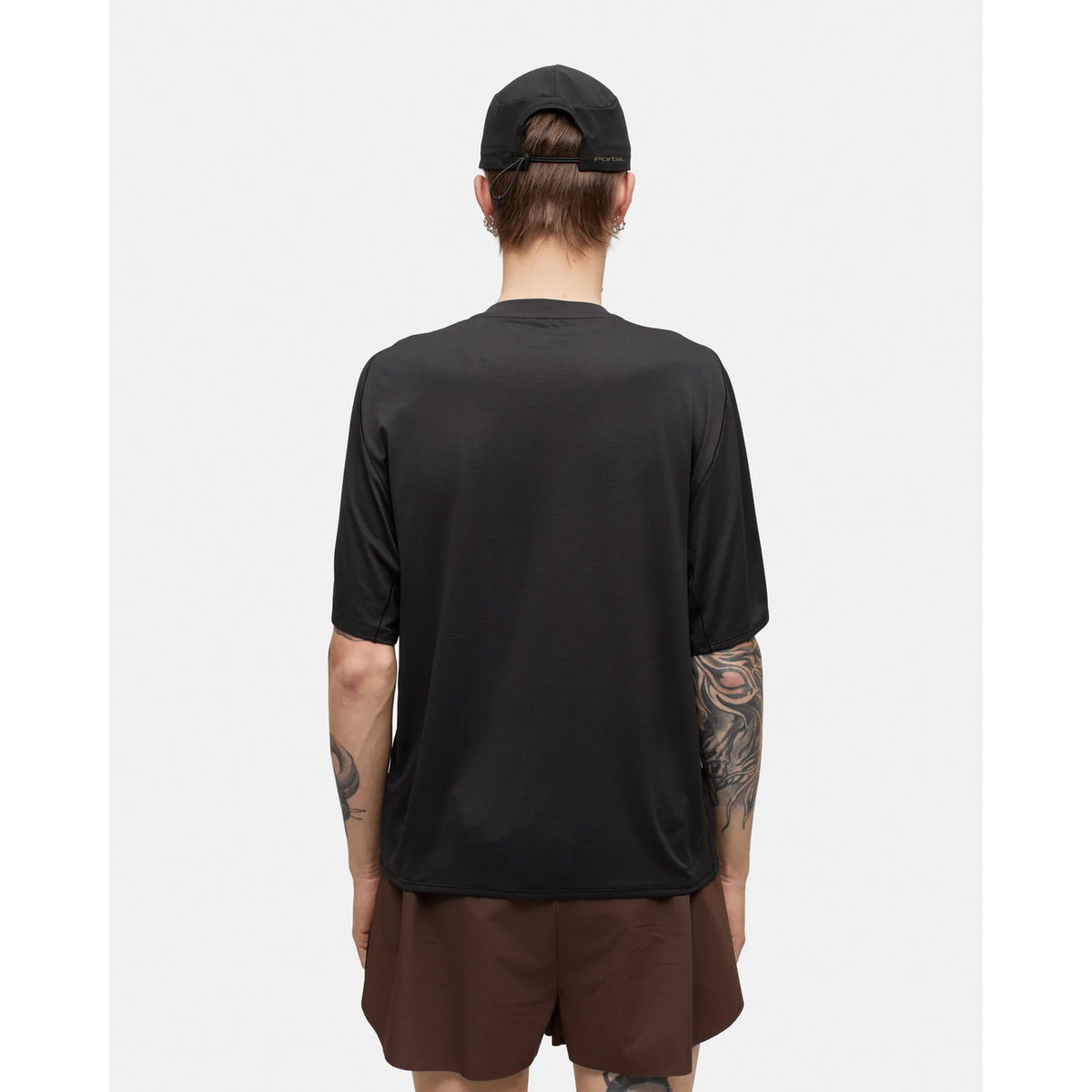 A tattooed individual wearing a black cap, brown shorts, and the Portal Men’s Lateral Peak Tee in Black Beauty—crafted from recycled polyester for moisture-wicking comfort—stands facing away from the camera against a plain white background.