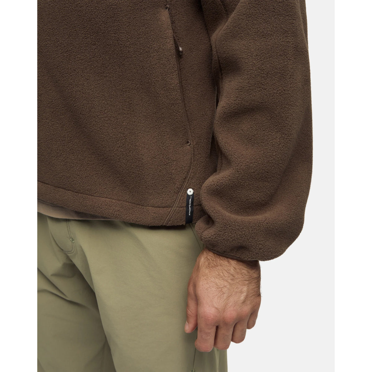 Close-up of a brown Portal Men’s Loam Fleece Jacket Slate Black with a small brand tag on the hem, worn over olive green pants.