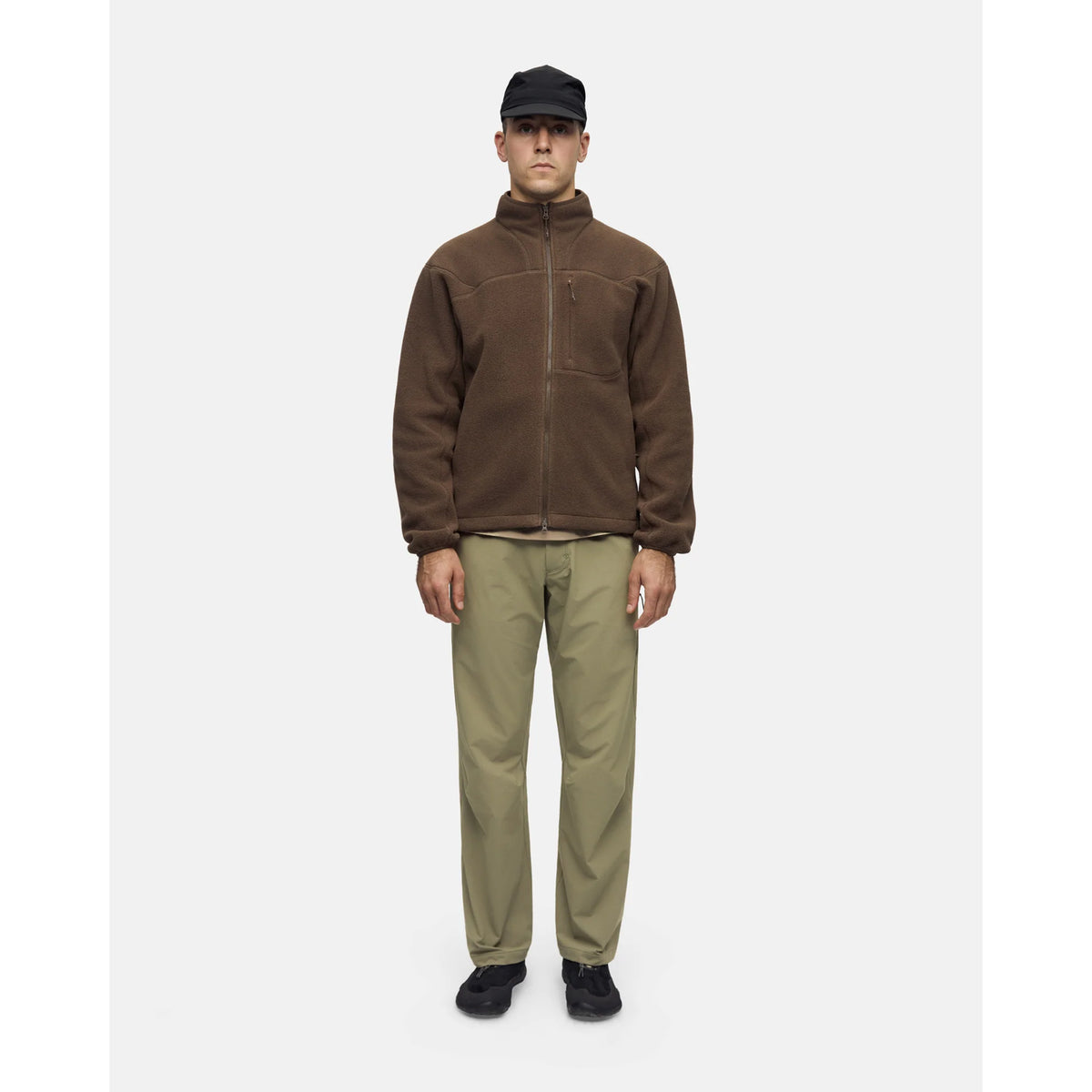 A person wearing a brown Men’s Loam Fleece Jacket Slate Black with a zippered chest pocket, khaki pants, a black baseball cap, and black sneakers.