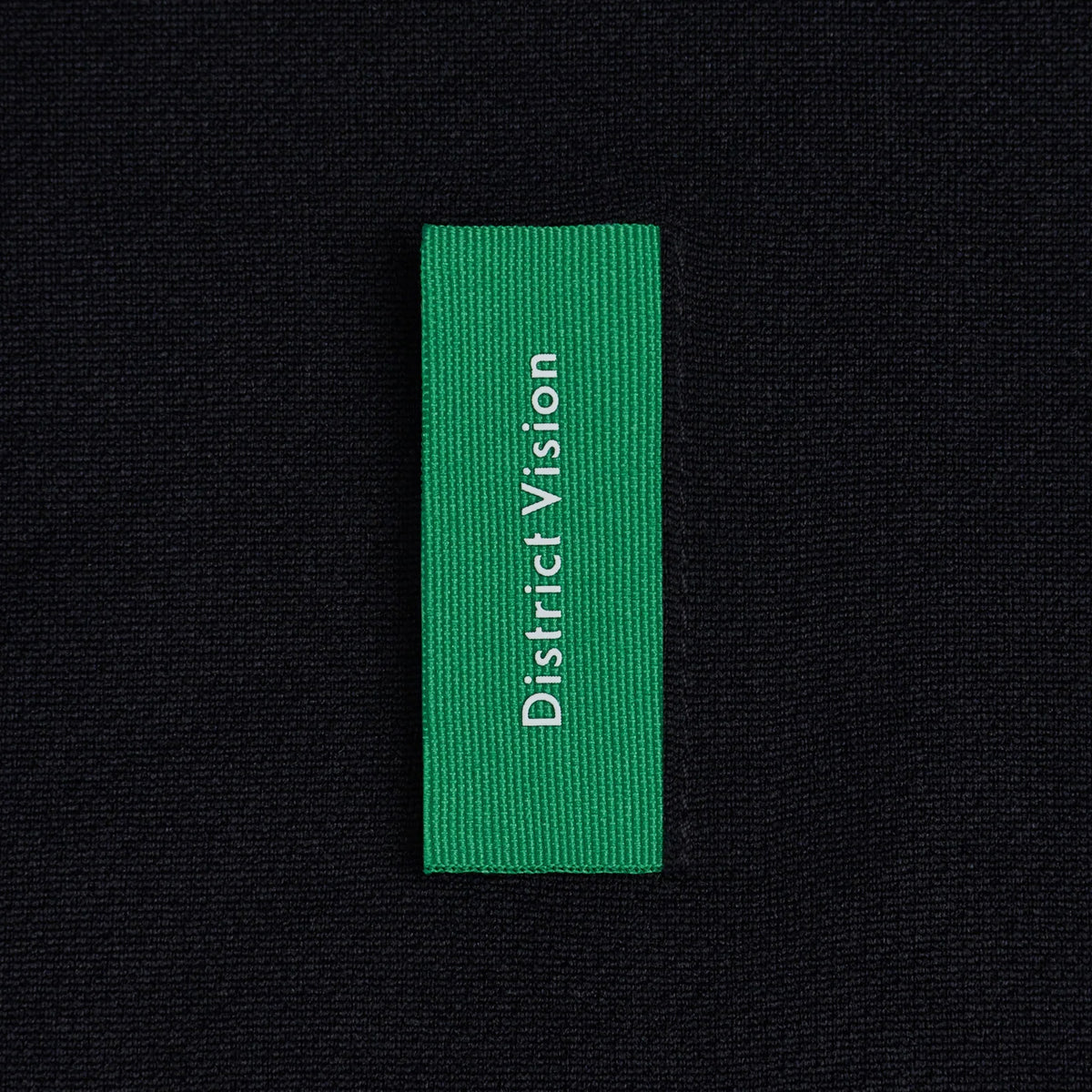A green rectangular District Vision fabric tag attached to the black, breathable stretch fabric of a Men’s Short Sleeve Boxy Training Tee Black Flame.