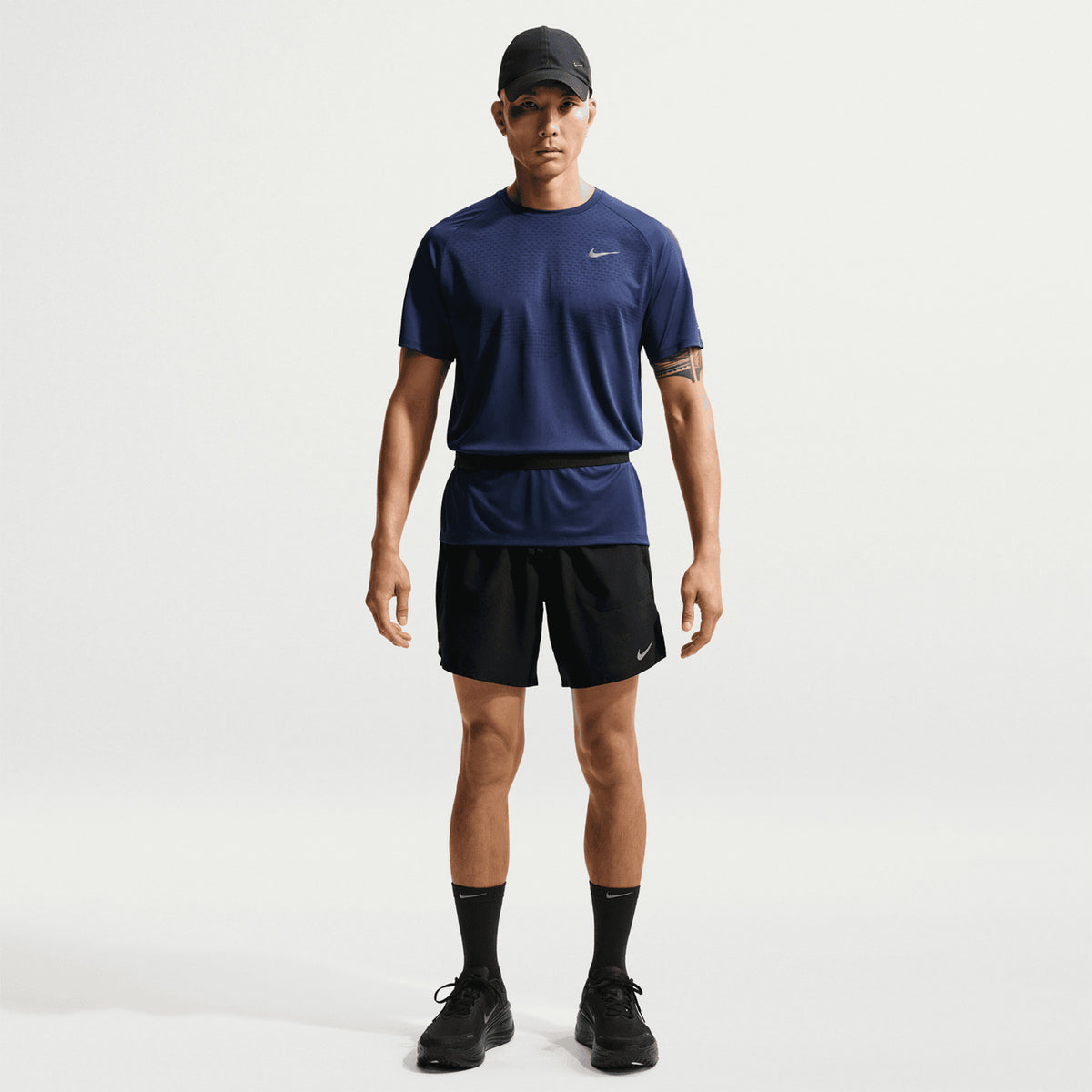 A person in a navy Nike T-shirt, black Nike Men’s Stride Dri-FIT 7 Inch Running Shorts featuring a zippered phone pocket, black cap, socks, and shoes stands facing forward against a plain white background.
