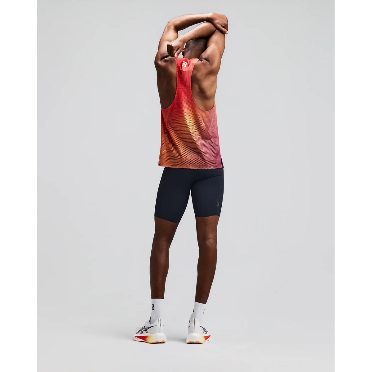 A person in a red tank top and black shorts stands facing away, stretching their arms overhead. They wear white socks and athletic shoes on a plain light background, with their athletic look completed by Bandit Men’s Vanisher Half Tights Anthracite—sweat-hiding tights that feature advanced Vanisher compression technology and a convenient seven-pocket system.