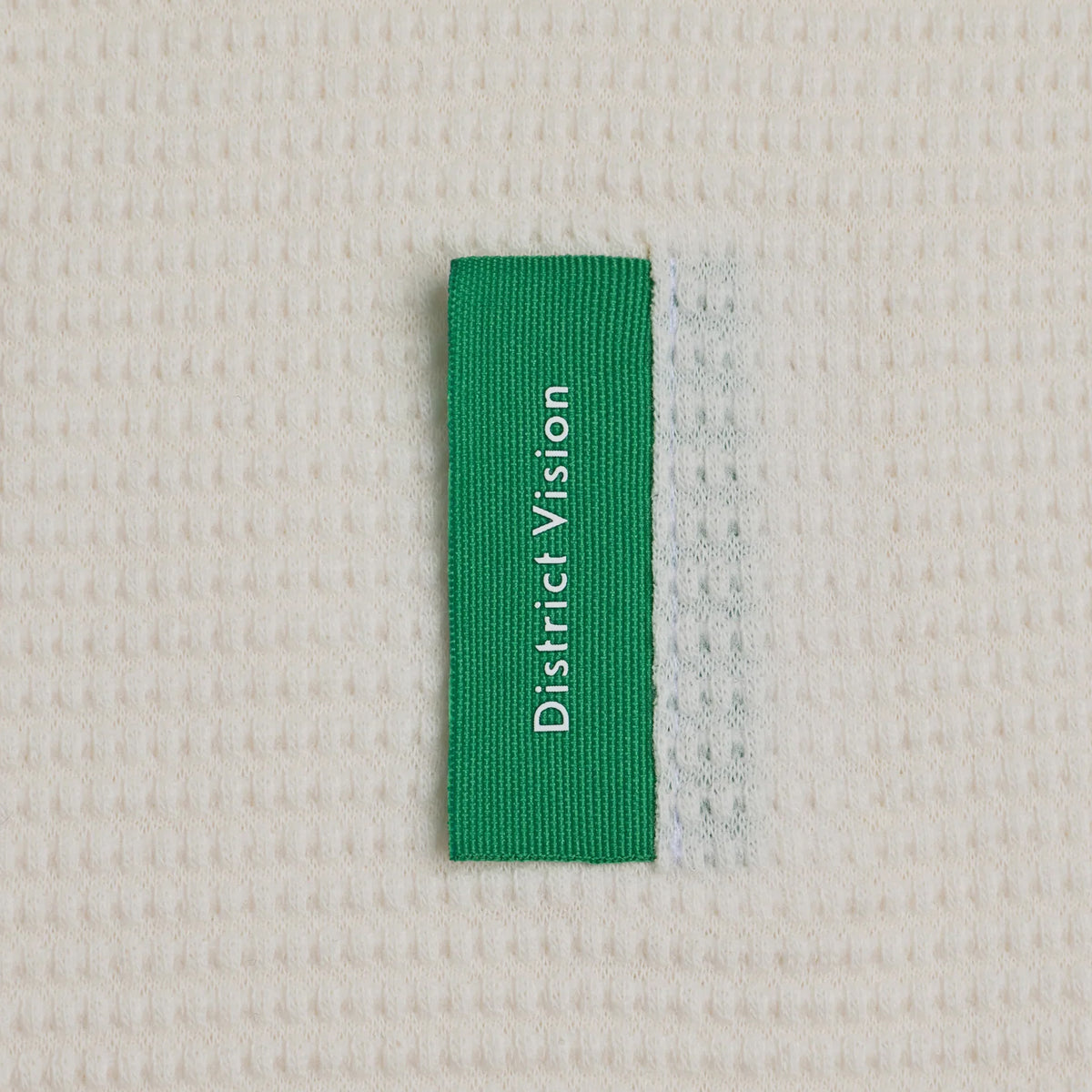 Green rectangular District Vision clothing label with the brand name in white text, sewn onto an off-white Men’s Waffle Top White long sleeve.