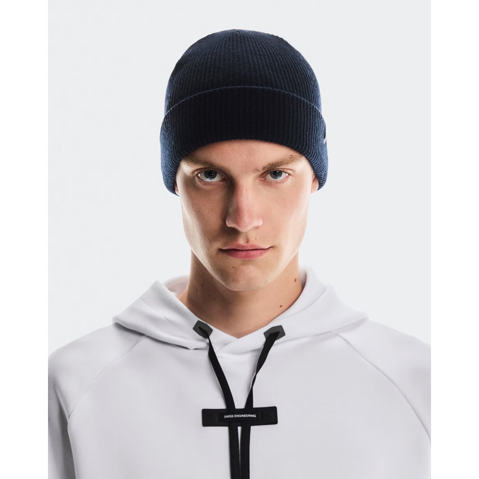 A person wearing the On Merino Beanie Midnight—a dark blue Merino wool beanie—paired with a white hoodie featuring black drawstrings, stands against a plain light background, looking directly at the camera with a neutral expression. The On Merino Beanie Midnight provides excellent temperature regulation for comfortable everyday wear.
