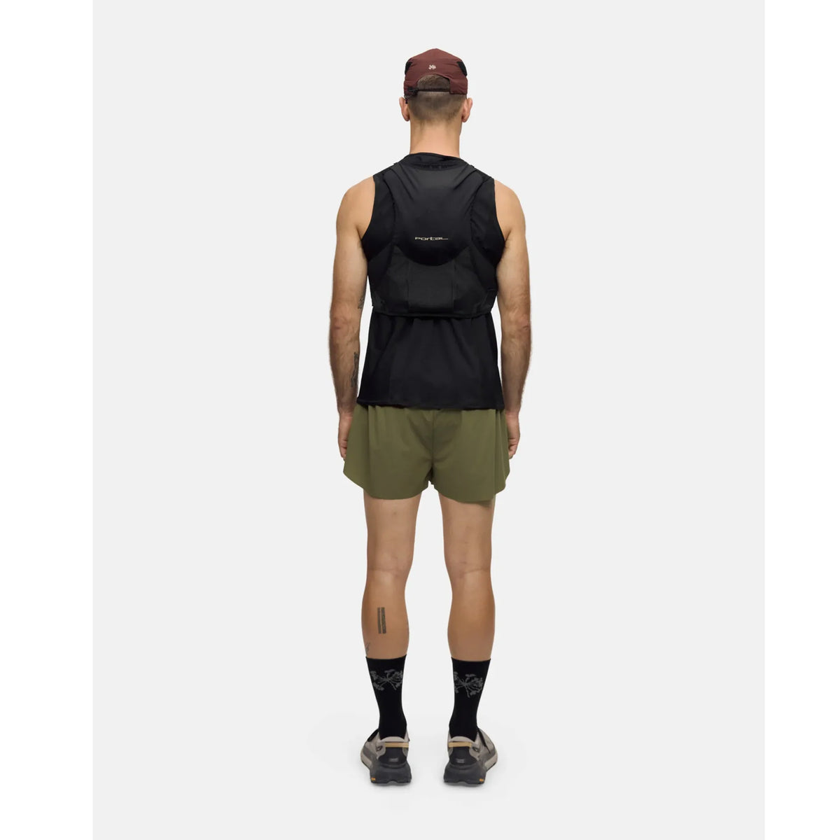 A man wearing a maroon cap, black sleeveless top, olive green shorts, black socks, and running shoes stands facing away from the camera against a plain light background, with the Portal Passage Cargo Vest Black Beauty slung over his shoulders.