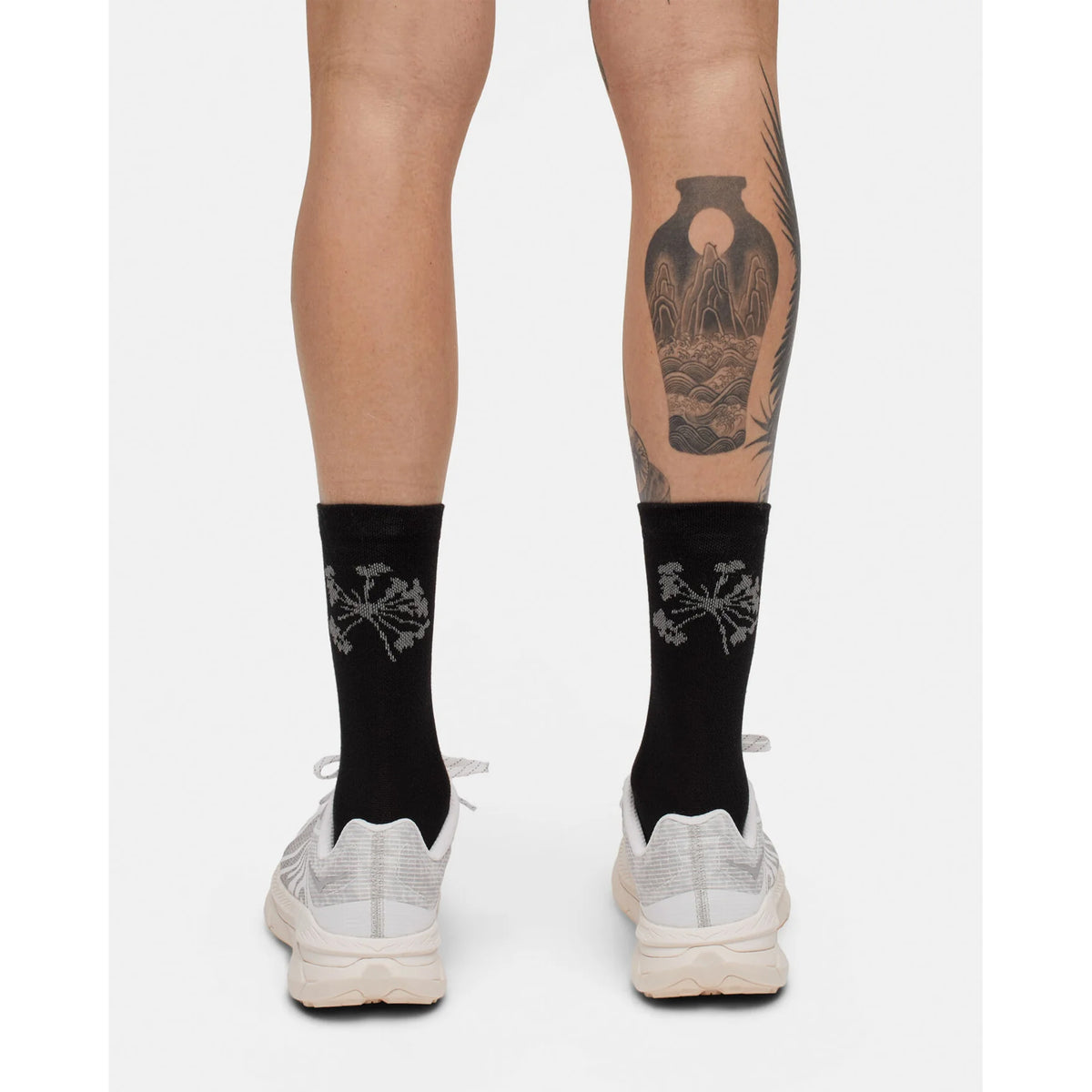 A person, seen from behind, is wearing Portal Pattern Active Socks Black Beauty / Reflective paired with white sneakers. The black socks feature a gray plant design, and one calf displays a prominent tattoo of a vase with decorative patterns. The background is plain white.