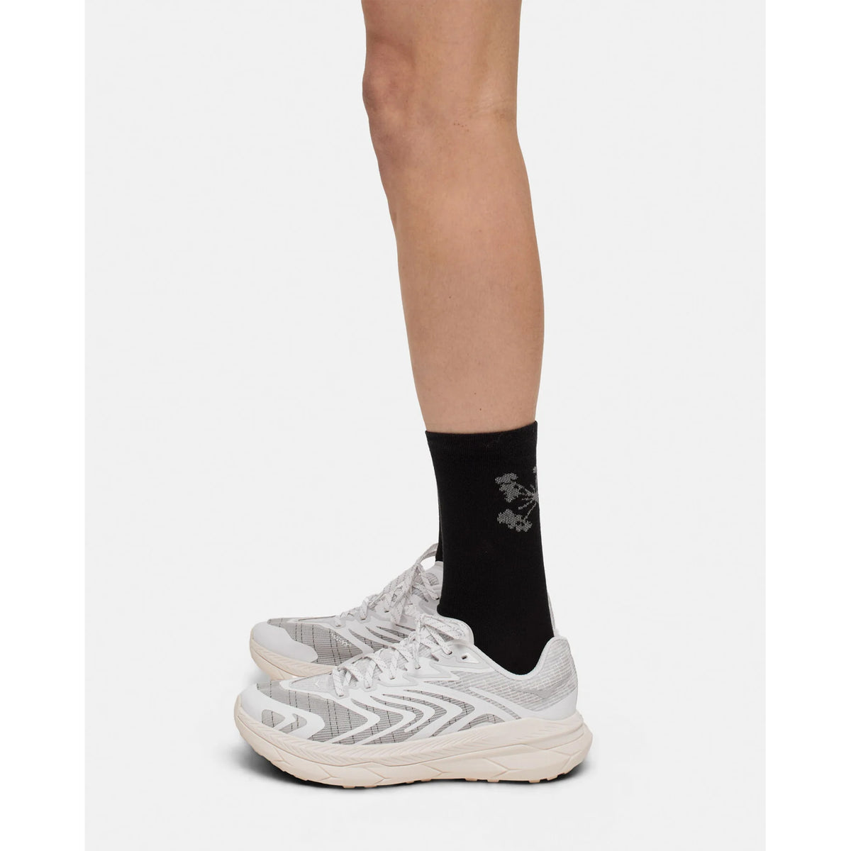 A person stands on a plain light background, shown from the side, mid-calf down, wearing Portal Pattern Active Socks in Black Beauty/Reflective—with a subtle design and moisture control properties—paired with white athletic shoes.