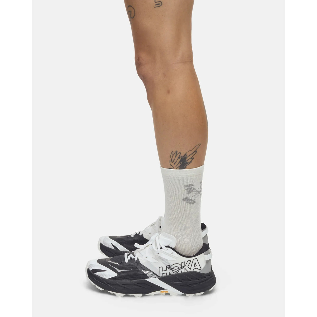 A person stands sideways against a plain white background, wearing Portal Pattern Active Socks in Bone White—lightweight synthetic socks designed for active use—paired with white and gray HOKA sneakers. Tattoos, including a bird and geometric shape, are visible on their leg.