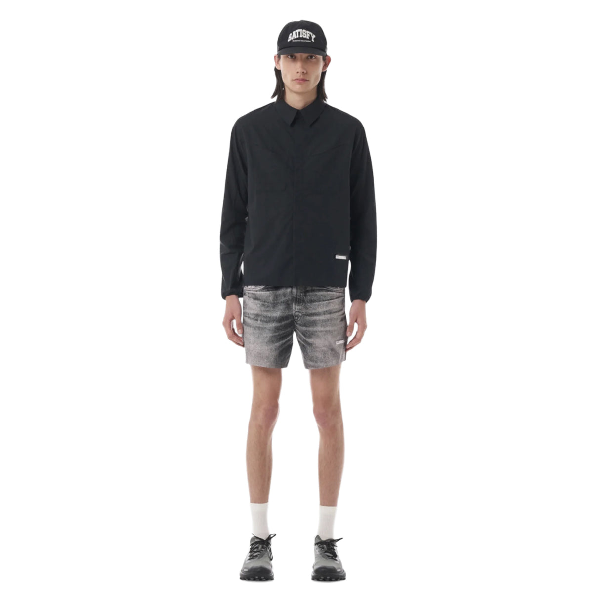 A person wearing a black cap, Satisfy PeaceShell River Shirt Black—a technical running shirt designed for lightweight wind protection—gray denim shorts, white socks, and black shoes stands against a plain white background.