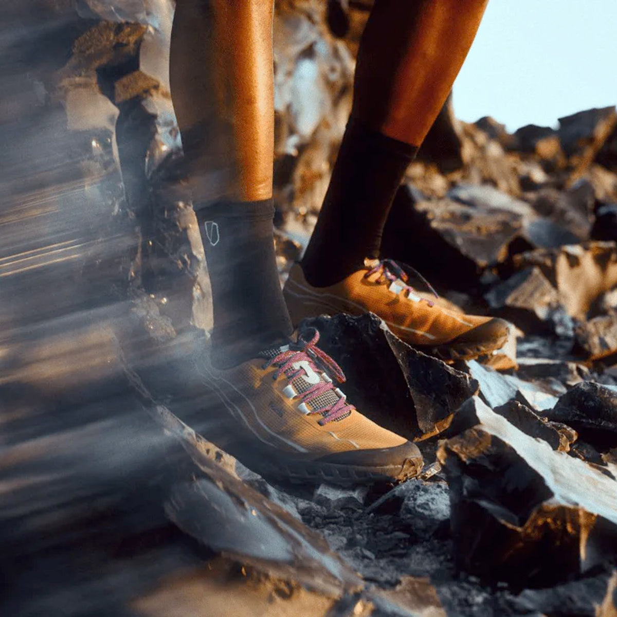 A person wearing black socks and tan trail running shoes stands on rocky terrain, with sunlight highlighting their legs and rocks around them.