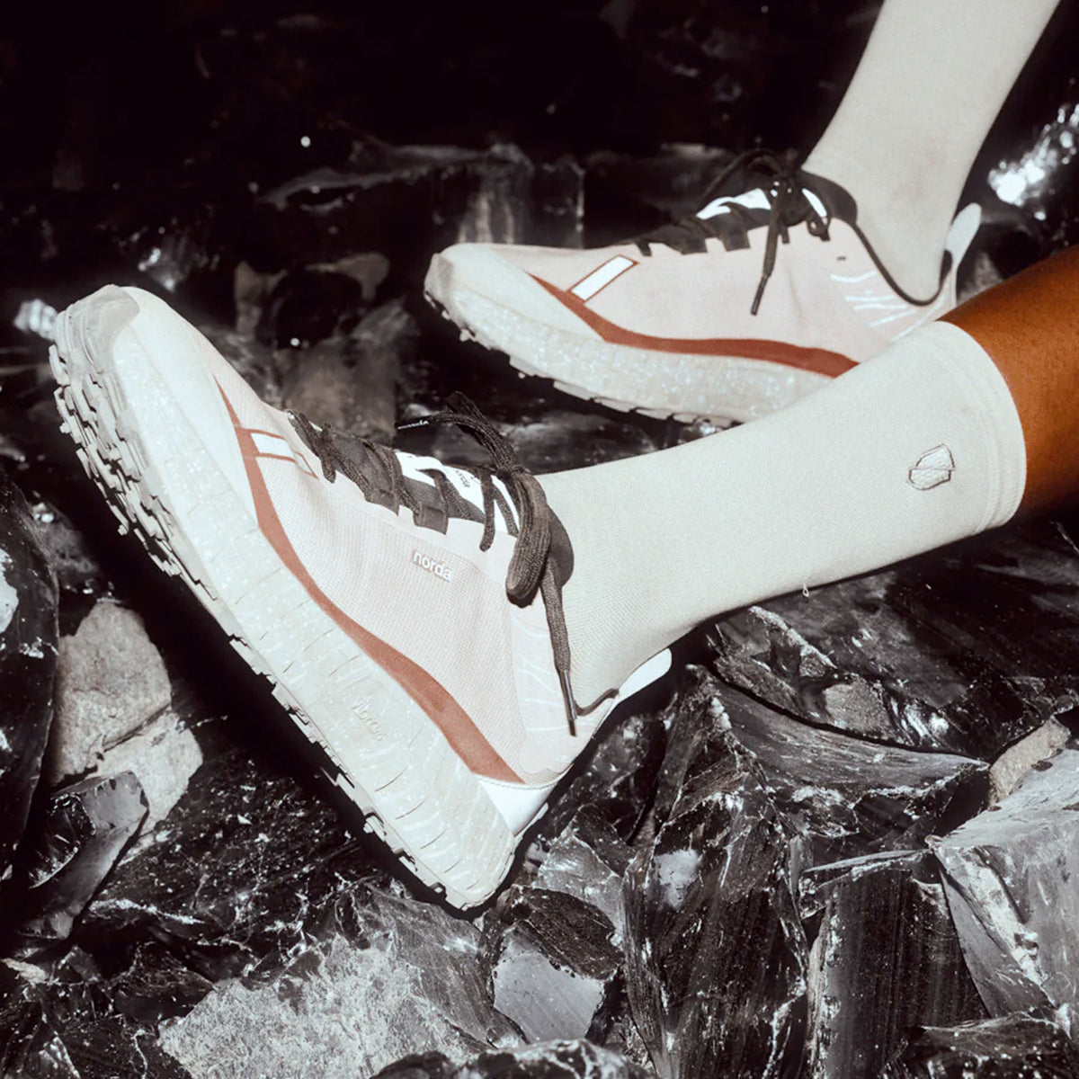 Close-up of a person wearing light pink and white sneakers with black laces and white socks, standing on a dark, reflective, rocky surface.