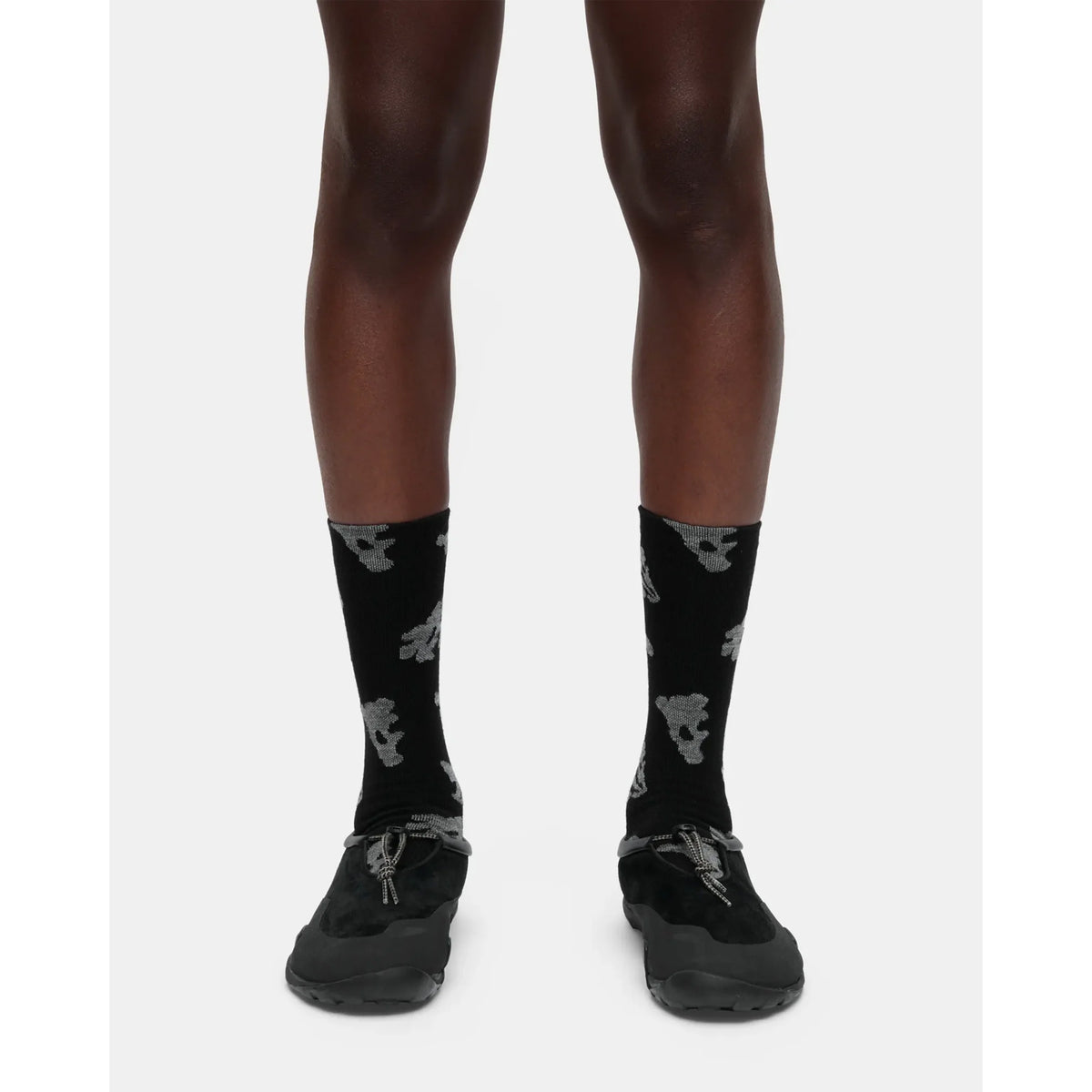 A person is wearing Portal Ritual Merino Socks Black Beauty, which feature a gray skull pattern, along with black shoes. The image shows only the lower legs and feet against a plain white background.