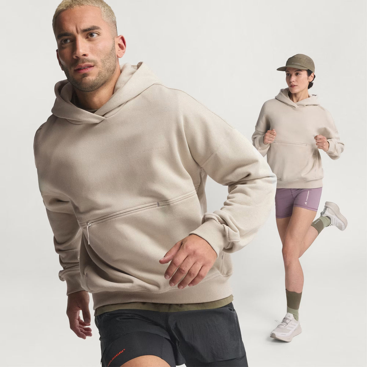A man and a woman are shown jogging against a plain white background, both wearing the Unisex Adi365 H.Koumori Running Hoodie in Putty Grey from Adidas. The woman pairs the hoodie with a cap, shorts, and socks, while the man styles it with black shorts.