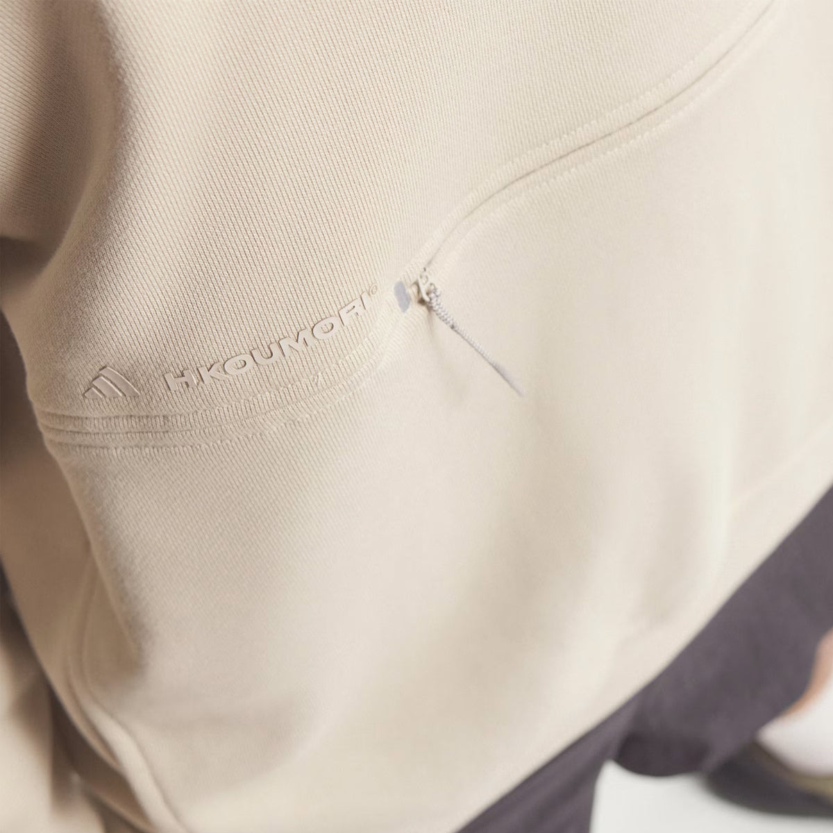 Close-up of the Unisex Adi365 H.Koumori Running Hoodie in Putty Grey by Adidas, showcasing a small zippered pocket and subtle embroidered branding on the left sleeve. Designed for all genders, this comfortable hoodie pairs seamlessly with dark pants, which are partially visible at the bottom.